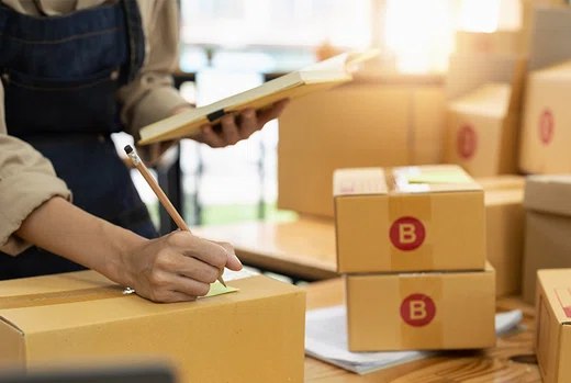Person holding a notebook and writing on a cardboard box, with more boxes marked with a red letter stacked behind.